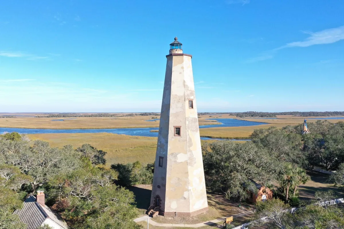 An iconic lighthouse rises above a backdrop of marshland and a river beneath a wide blue sky. The area around the lighthouse is open and inviting, with people potentially enjoying picnics or taking photographs, evoking a sense of adventure and exploration.