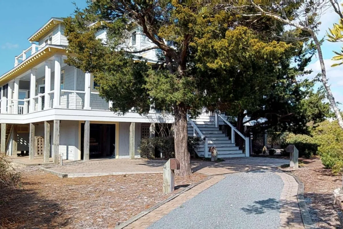 A large, welcoming house sits amid lush trees, with a stone pathway leading to the entrance and a cozy porch ideal for outdoor relaxation. The surrounding greenery and shaded setting evoke a tranquil retreat close to nature and trails.