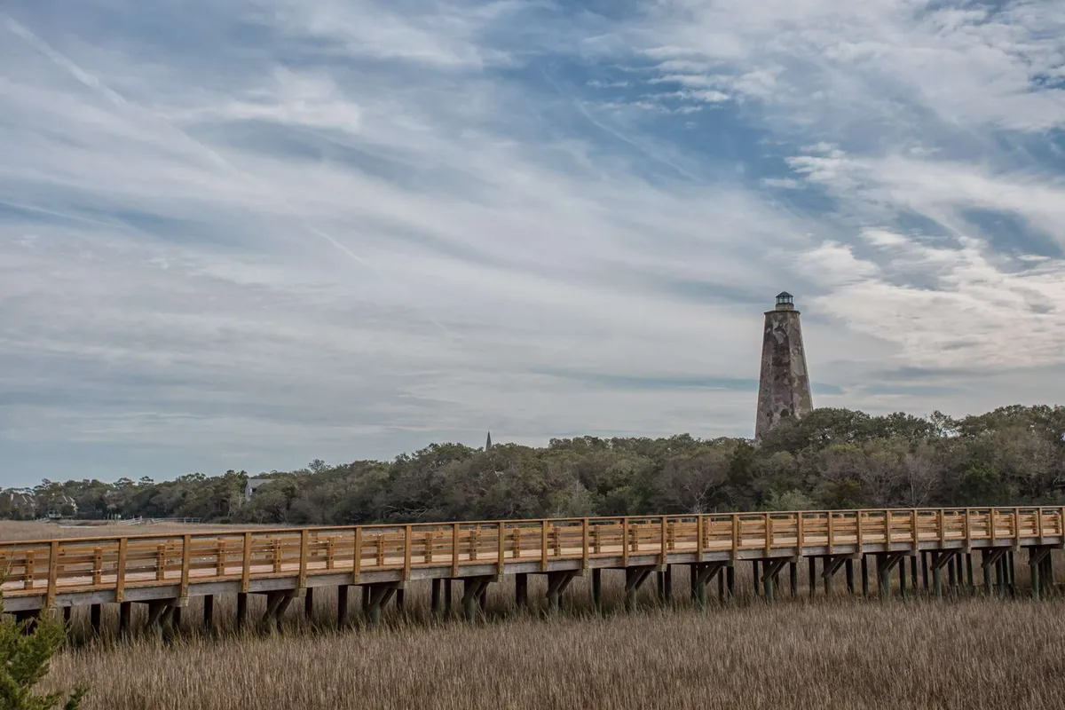 A scenic exterior view featuring a wooden boardwalk leading to the iconic lighthouse, with marshland and a cloudy sky in the background. The setting is tranquil, inviting exploration and nature walks.