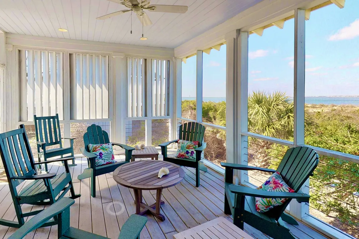 A cozy, screened-in porch furnished with rocking chairs, a central table, and tropical cushions. Large windows bring in natural light and frame the scenic outdoor views, creating a serene nook for reading, socializing, or enjoying the breeze without bugs.