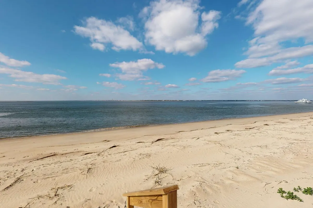 A serene sandy beach stretches along calm waters under a soft, cloudy sky. A distant yacht floats peacefully offshore, offering a sense of relaxation and leisurely island living.