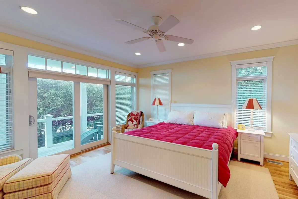 A serene bedroom featuring a plush bed with a red comforter, large windows that allow sunlight to fill the space, a ceiling fan, and side lamps. The room exudes warmth and comfort, with tranquil décor and an inviting ambiance. The large windows suggest a connection to the outdoors, possibly with access to a deck.