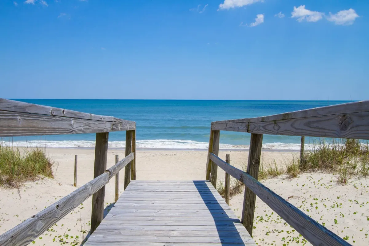 A wide, open view from a wooden walkway looking out onto a sandy beach, ocean waves, and lush dune grass. The perspective emphasizes the close proximity to the water and the inviting natural environment, suggesting easy, direct transitions from the home to the beach for swimming, sunbathing, or collecting shells.