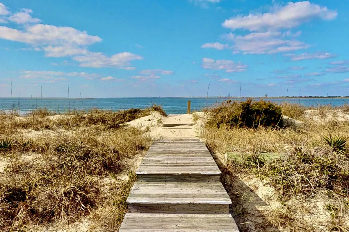 A serene exterior scene featuring a wooden walkway that leads directly over sand dunes to the ocean. Lush vegetation flanks the path, and the open view of the sea promises immediate access to the beach. The overall atmosphere is peaceful, inviting spontaneous beach activities such as walking, sandcastle building, and seashell collecting.