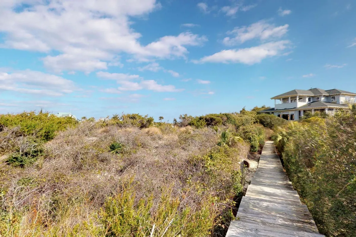 A serene exterior scene with a wooden walkway stretching through lush island vegetation, leading towards a distant beach house. The natural setting evokes outdoor activity and relaxation.