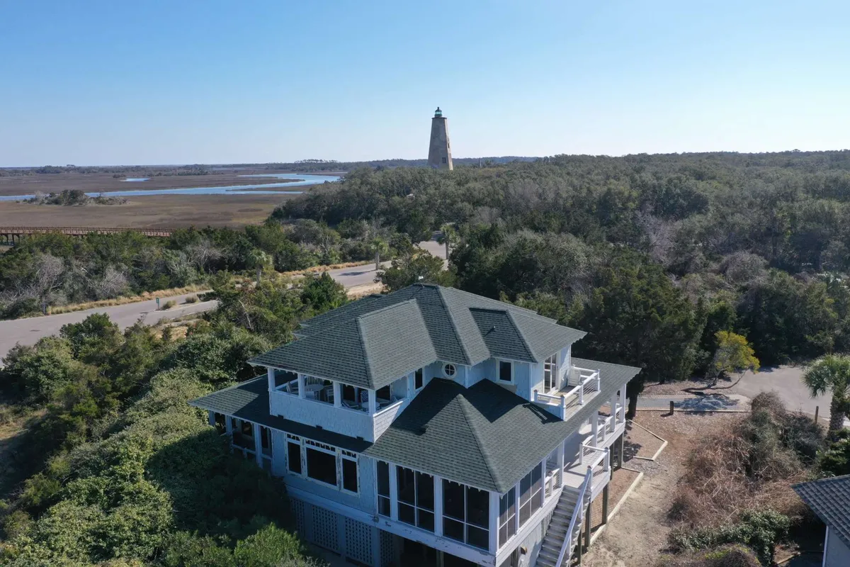An exterior view featuring the house's distinctive architecture, with the lighthouse visible nearby, surrounded by greenery and a nearby road. The peaceful setting underscores opportunities for watching the sunrise, stargazing, and taking in the broader island context.
