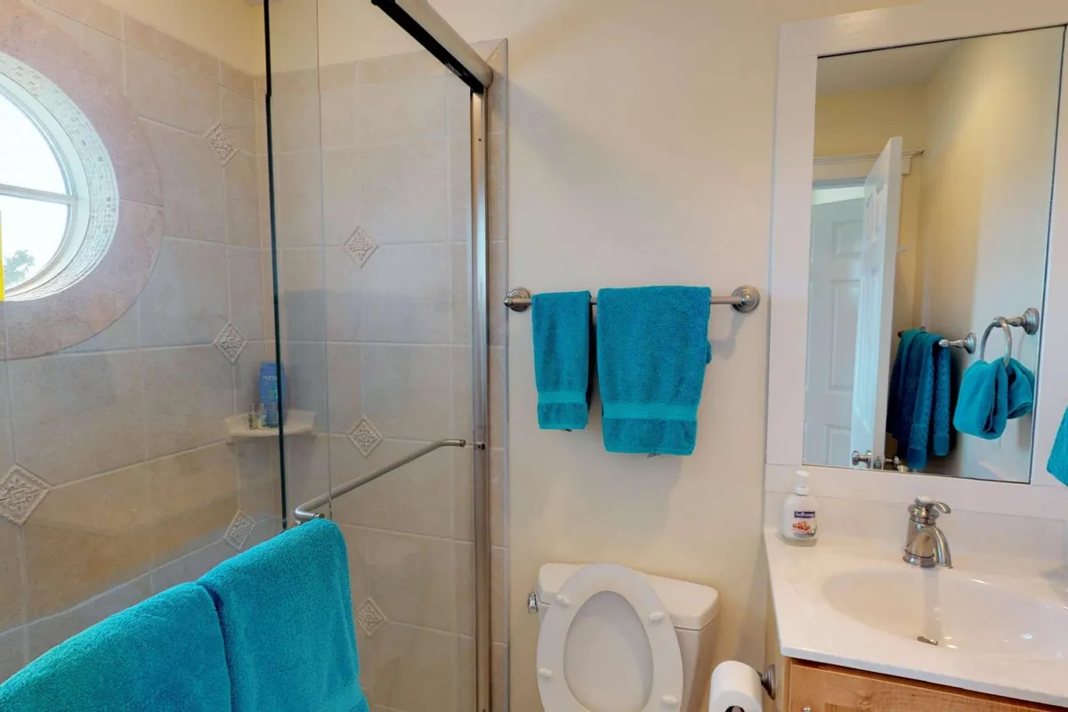 A serene bathroom with a glass-door shower, a round window, a sink area with mirror, and plush blue towels. The space is bright, inviting, and designed for relaxation and privacy.