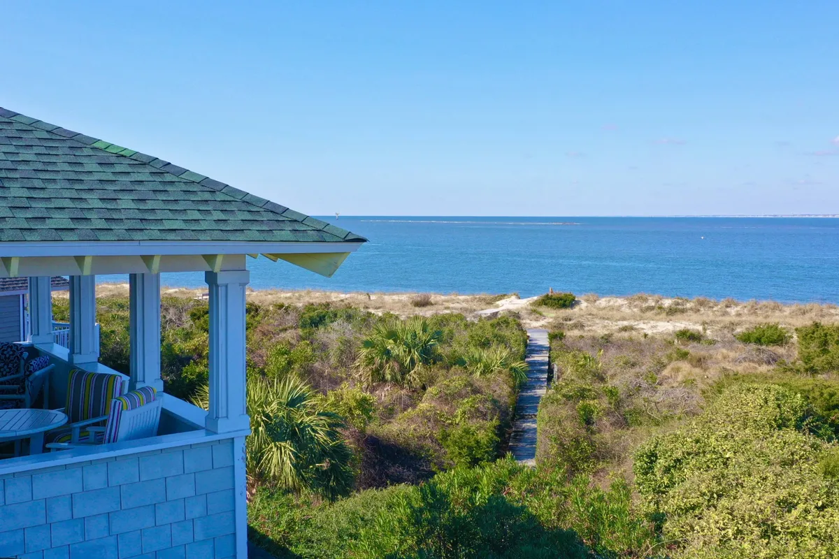 The image shows the exterior of a charming coastal home with a wooden pathway leading toward a wide, shaded porch. Beyond, there are sweeping ocean views, soft dunes, and hints of sunrise light. The scene evokes peaceful mornings, easy beach access, and a welcoming, relaxed atmosphere perfect for families and friends.