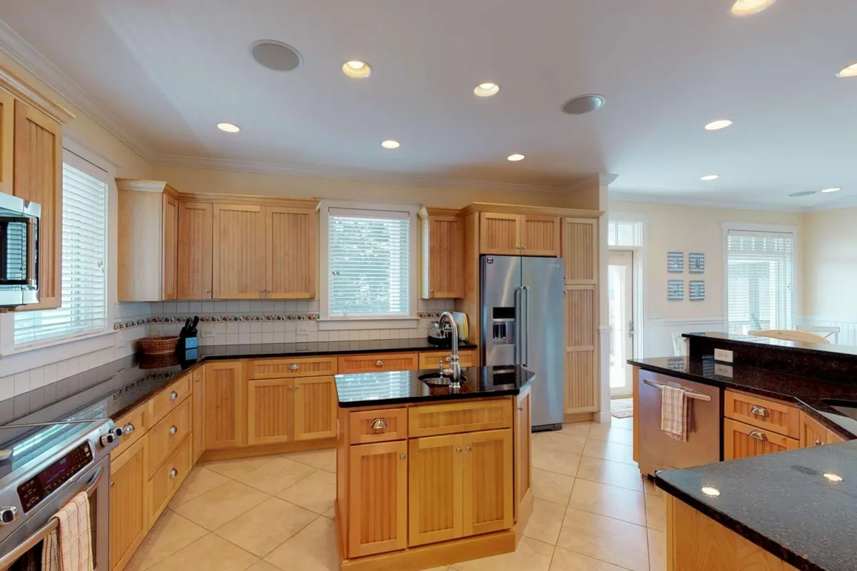 A modern kitchen features granite countertops, wooden cabinets, stainless steel appliances, and large sunlit windows. The layout is open and perfect for meal prep, entertaining, and family gatherings.