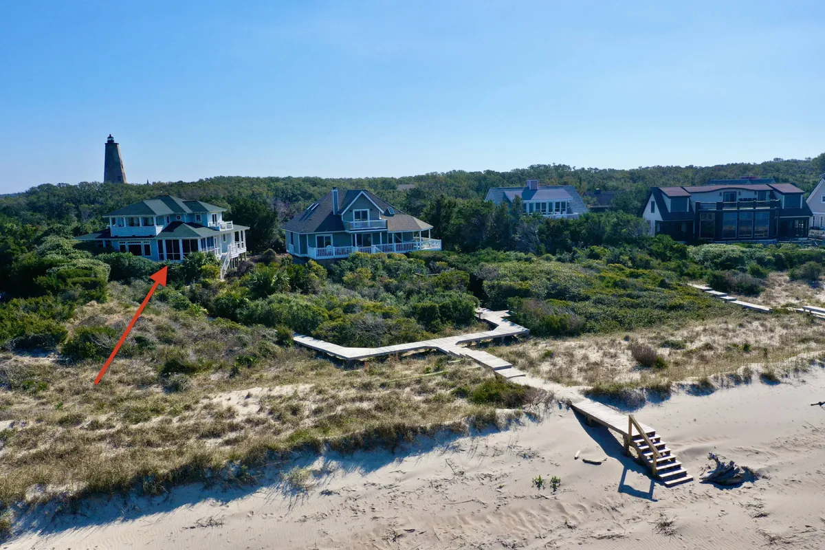 A relaxed exterior shot showcasing a beachfront house with a wooden pathway, lush greenery, and a distant lighthouse. The house is positioned right by the water, creating a tranquil, inviting setting ideal for family gatherings and spontaneous outdoor fun, as well as moments of quiet relaxation.