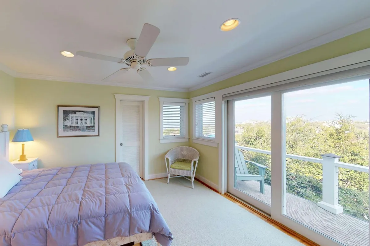 A tranquil bedroom with a bed dressed in light purple bedding, a blue lamp on the bedside table, and large windows that overlook a beautiful view. The natural light streaming in and the view suggest an inviting space ideal for relaxation, with possible access to an outdoor balcony or deck.