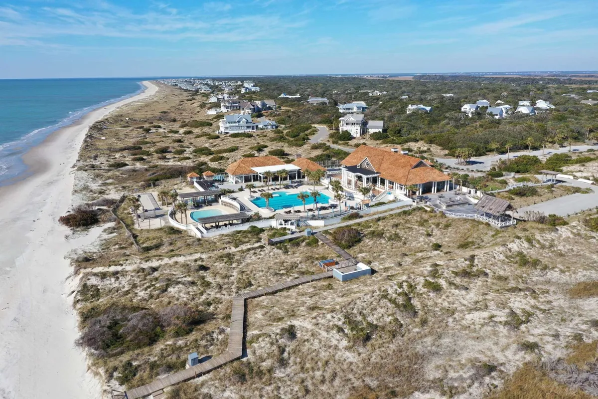 An expansive pool area stretches out towards the beachfront, with inviting lounge chairs and umbrellas. The ocean is visible just beyond the pool, and the surrounding landscape includes lush greenery and a glimpse of residential homes in the background. The mood is relaxed and inviting, suggestive of resort-style amenities and upscale leisure.